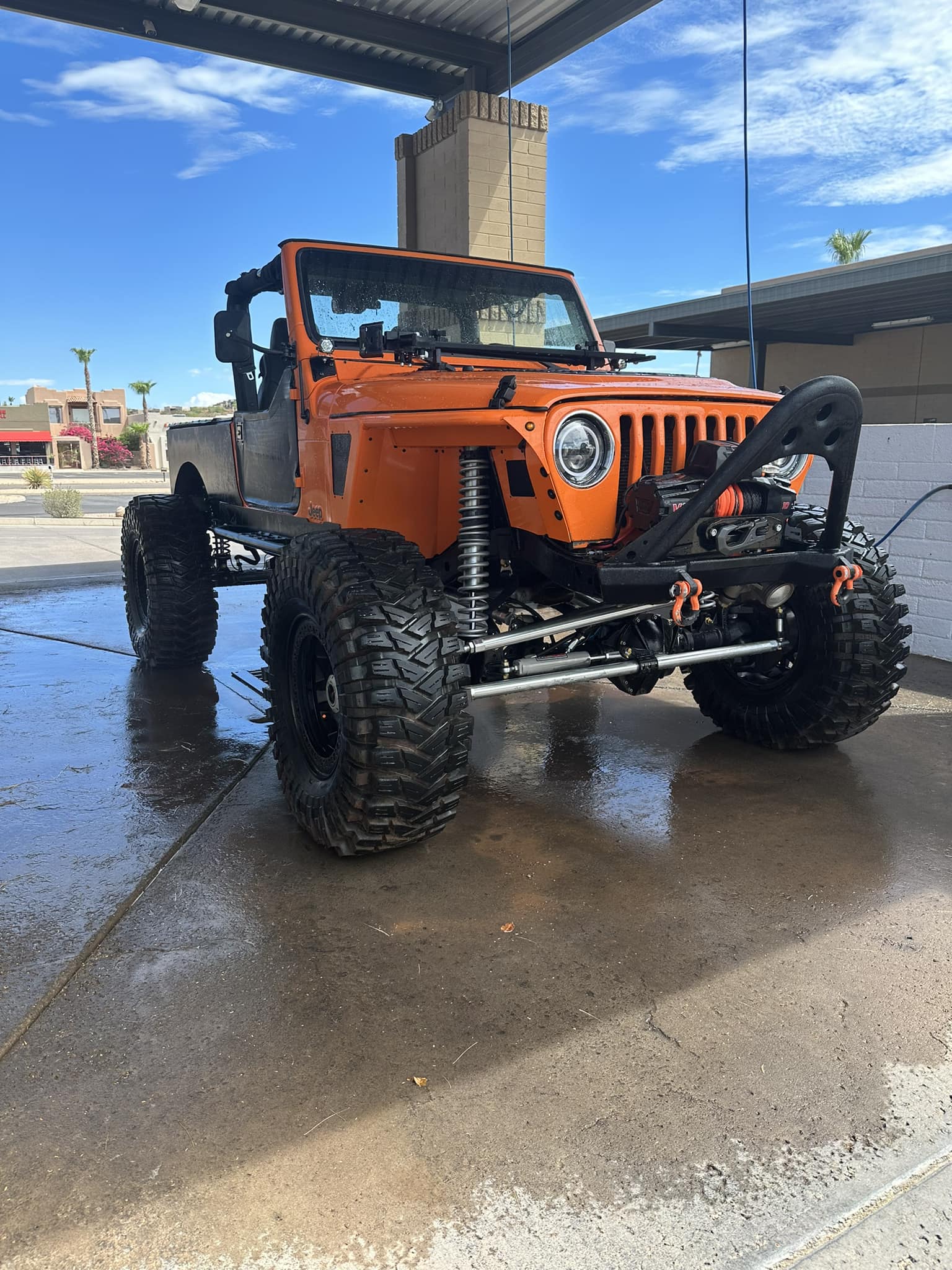Custom orange Jeep with oversized knobby tires and lifted suspension parked on wet concrete.