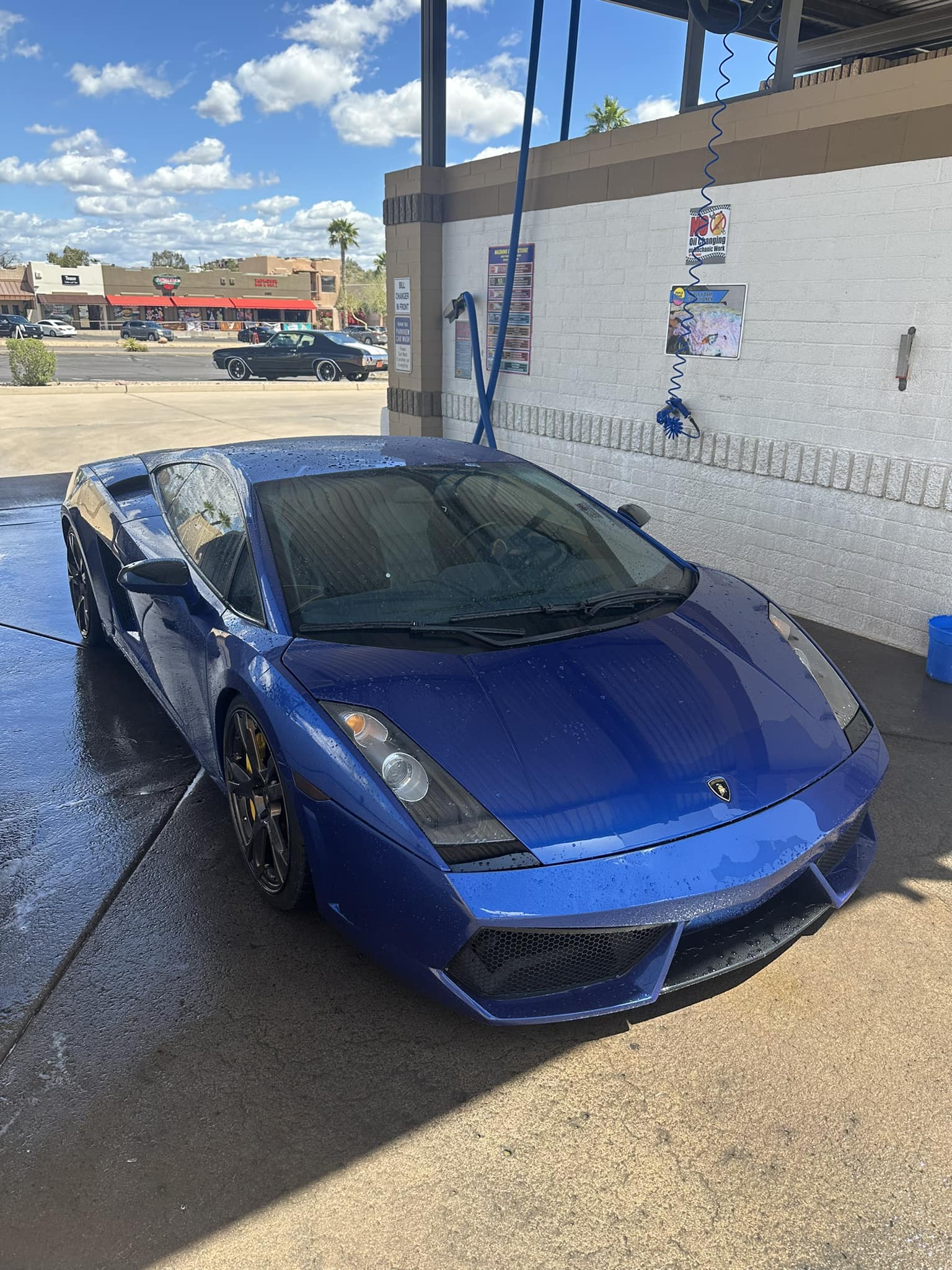 Wet blue Lamborghini Gallardo parked in a self-service car wash bay on a sunny day.